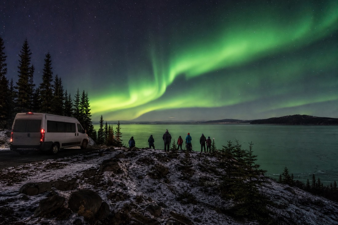 Small tour van waiting at a dark autumn viewpoint while guests watch the aurora over an open lake