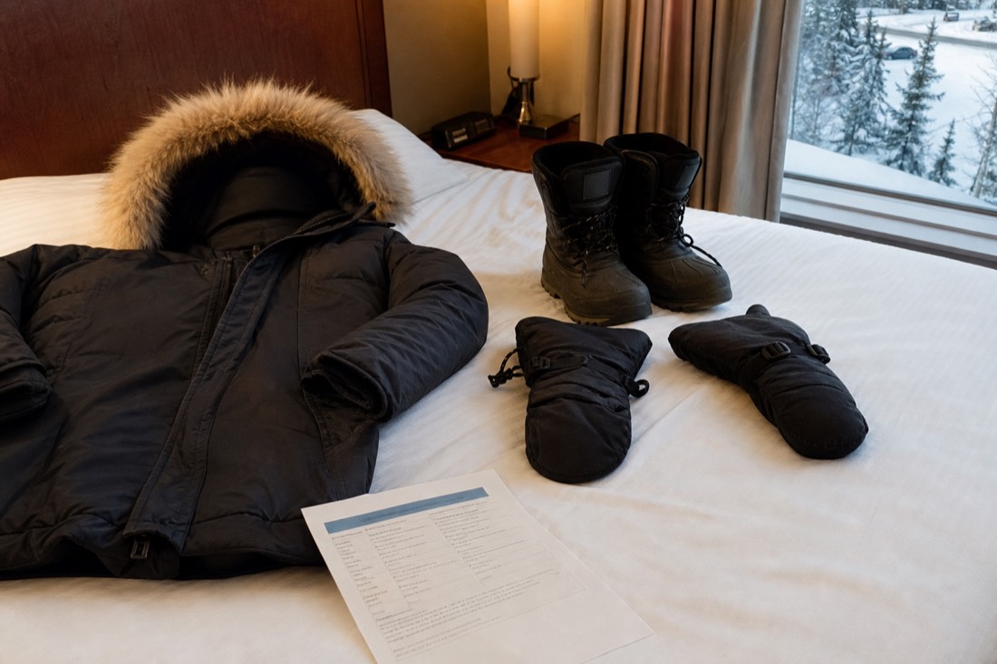 Rented arctic parka, insulated boots and mitts laid out on a hotel bed in Yellowknife beside a printed tour voucher