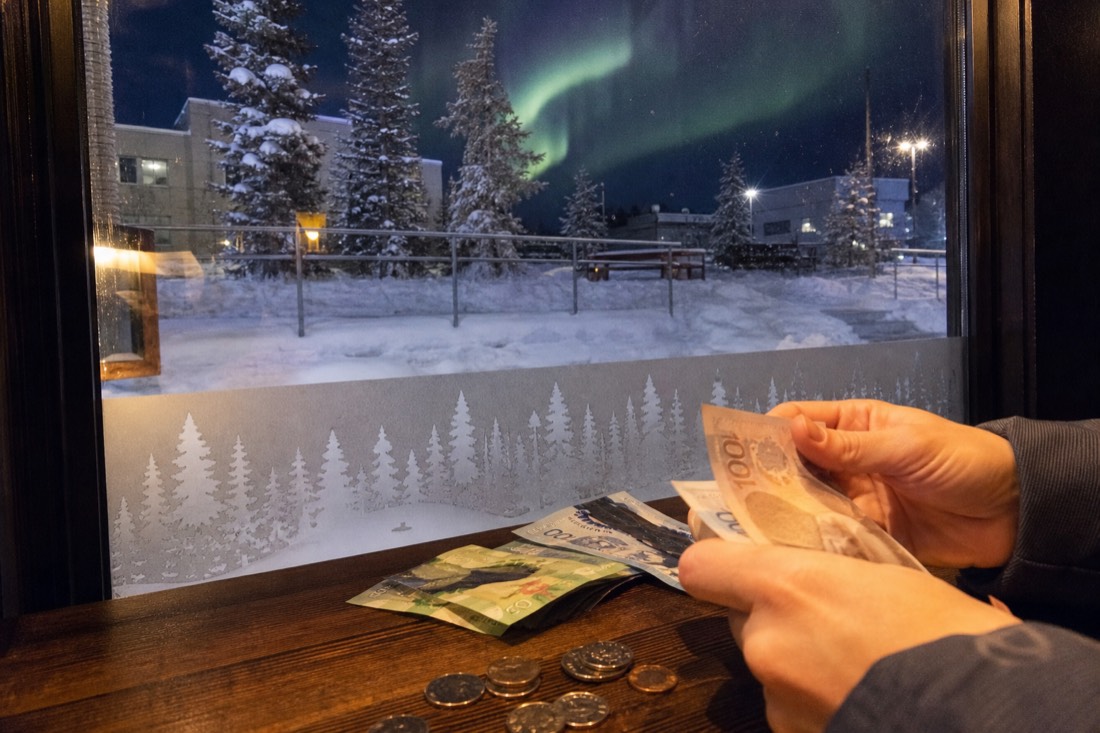Hands counting Canadian banknotes and coins on a cafe table in Yellowknife with a frosted window and snow outside