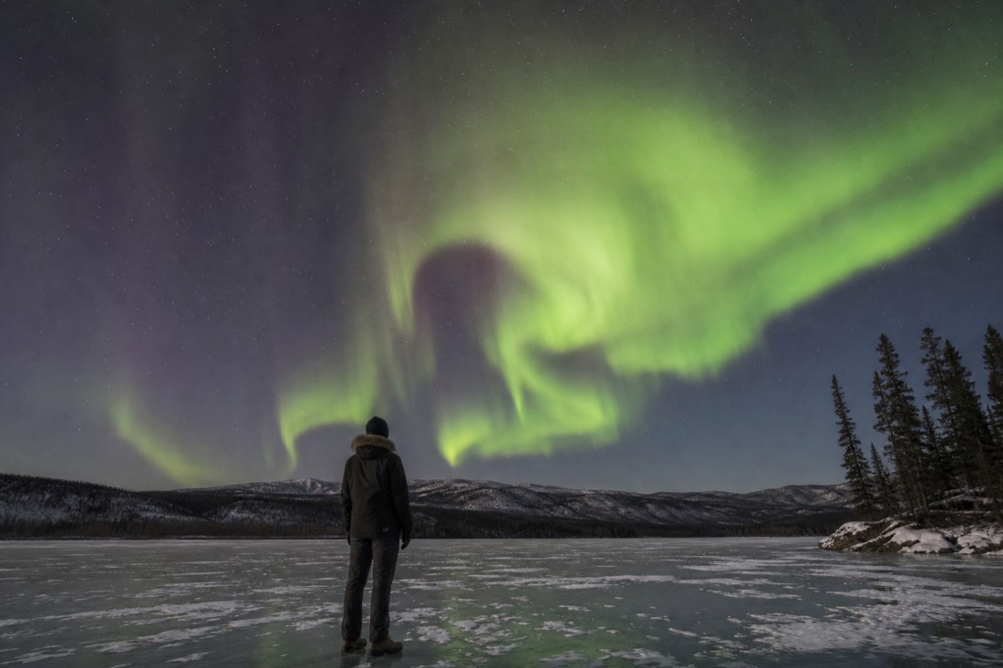 Traveler standing on a frozen lake at night looking up at a green aurora spiral overhead